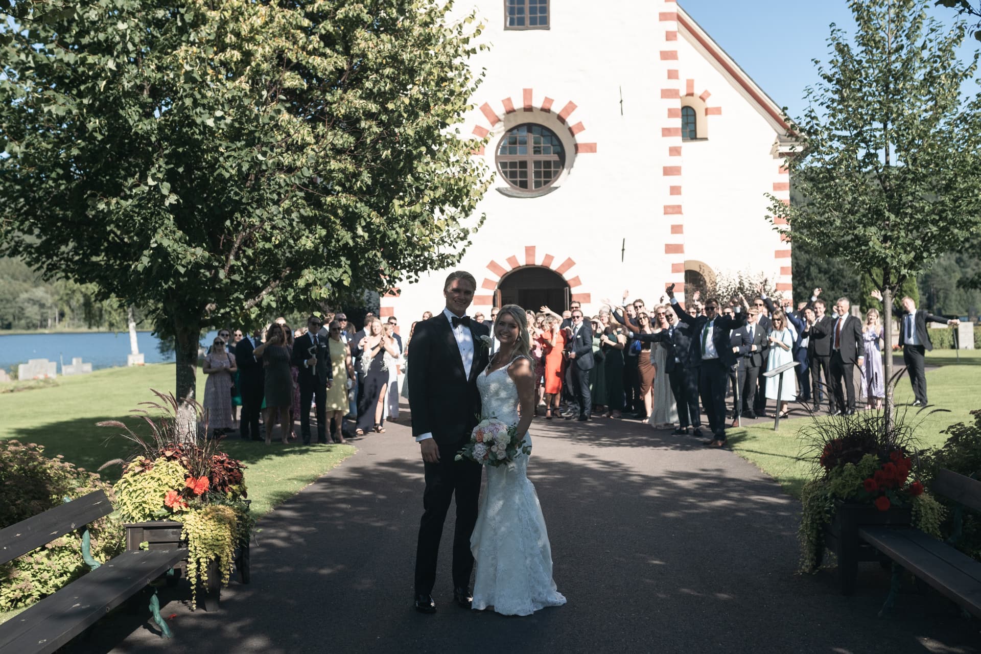Brudparet framför kyrkan medan gästerna jublar bakom dem – bröllopsfoto av Mehmet Budak