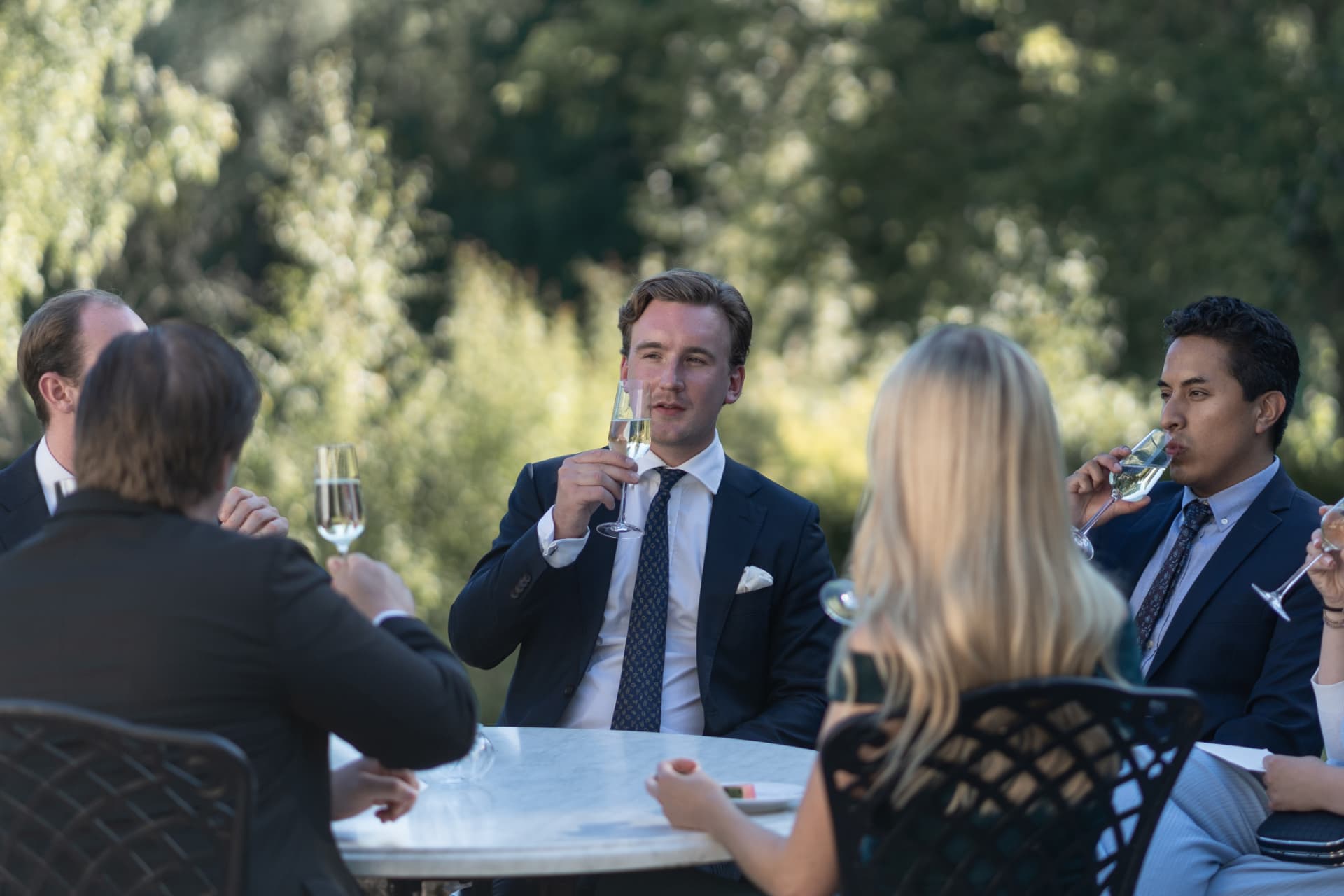 Gäster skålar i champagne runt ett bord i herrgårdens trädgård under mottagningen – bröllopsfoto av Mehmet Budak