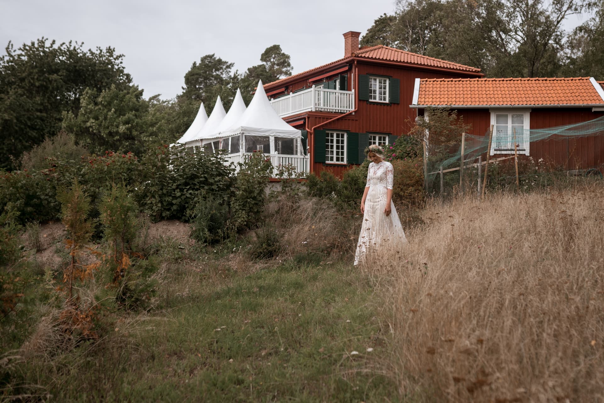 Bruden går i ängen med festtältet och röda hus bakom – bröllopsfoto av Mehmet Budak