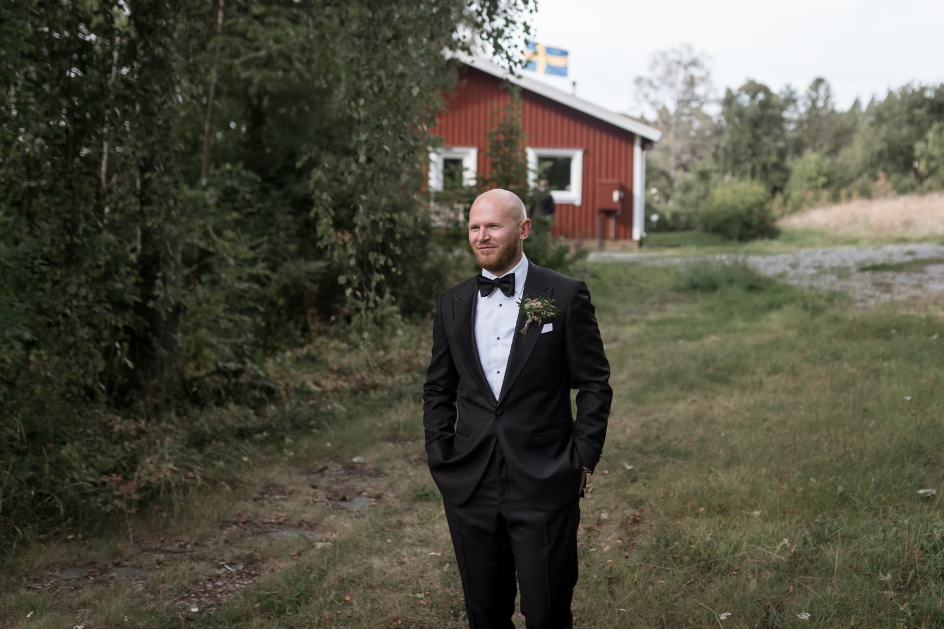 Brudgummen i smoking framför en röd stuga med svensk flagga – bröllopsfoto av Mehmet Budak