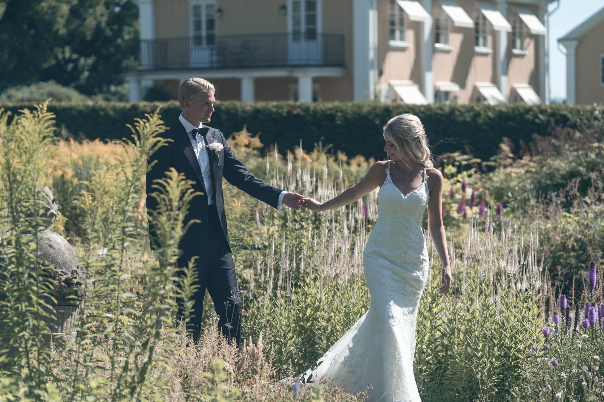 Brudparet går hand i hand genom parken med Ulvsunda Herrgård bakom sig – bröllopsfoto av Mehmet Budak