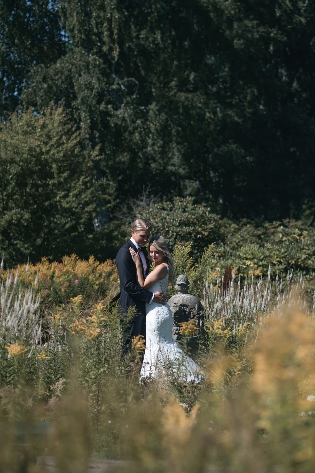 Brudparet står nära varandra vid stenurnan bland höga blommor – bröllopsfoto av Mehmet Budak