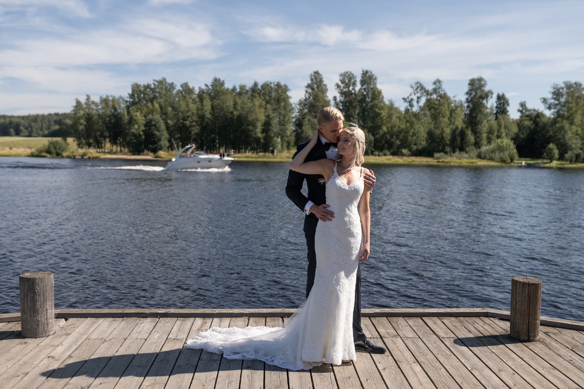 Brudparet på bryggan med en båt som passerar i bakgrunden – bröllopsfoto av Mehmet Budak