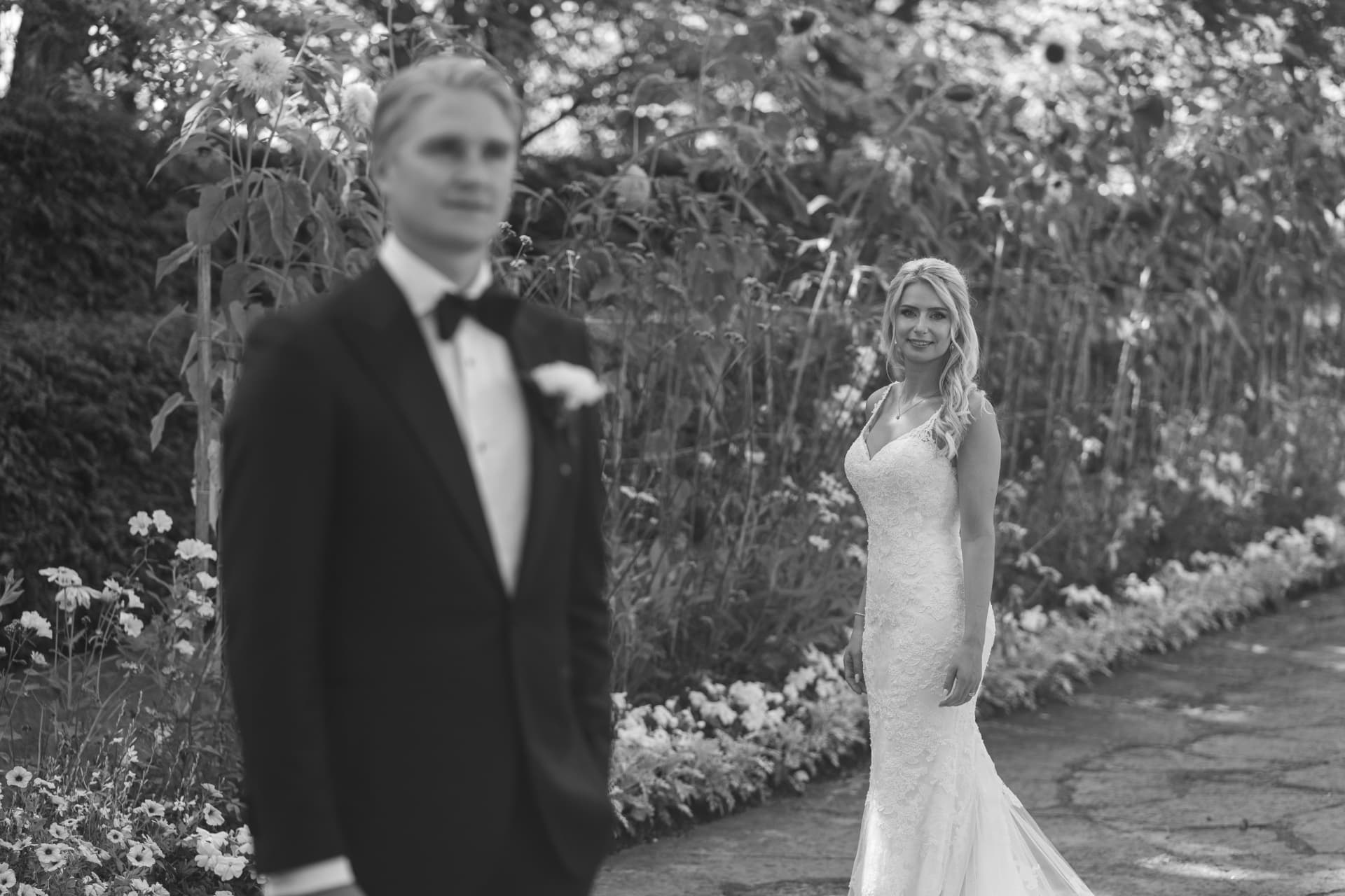 Bruden ler mot kameran bakom brudgummen i blomsterrabatten, svartvitt – bröllopsfoto av Mehmet Budak