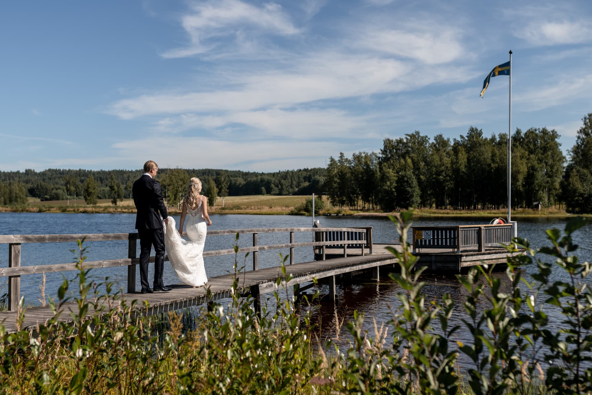 Brudparet promenerar ut på bryggan med svenska flaggan vid sjön – bröllopsfoto av Mehmet Budak