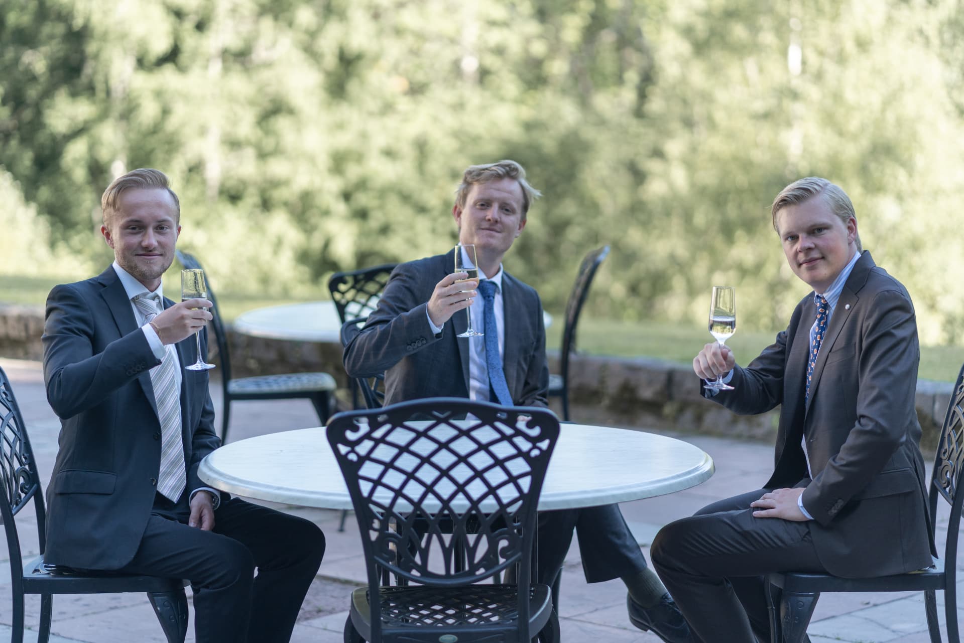 Tre gäster i kostym höjer sina champagneglas vid ett bord på terrassen – bröllopsfoto av Mehmet Budak
