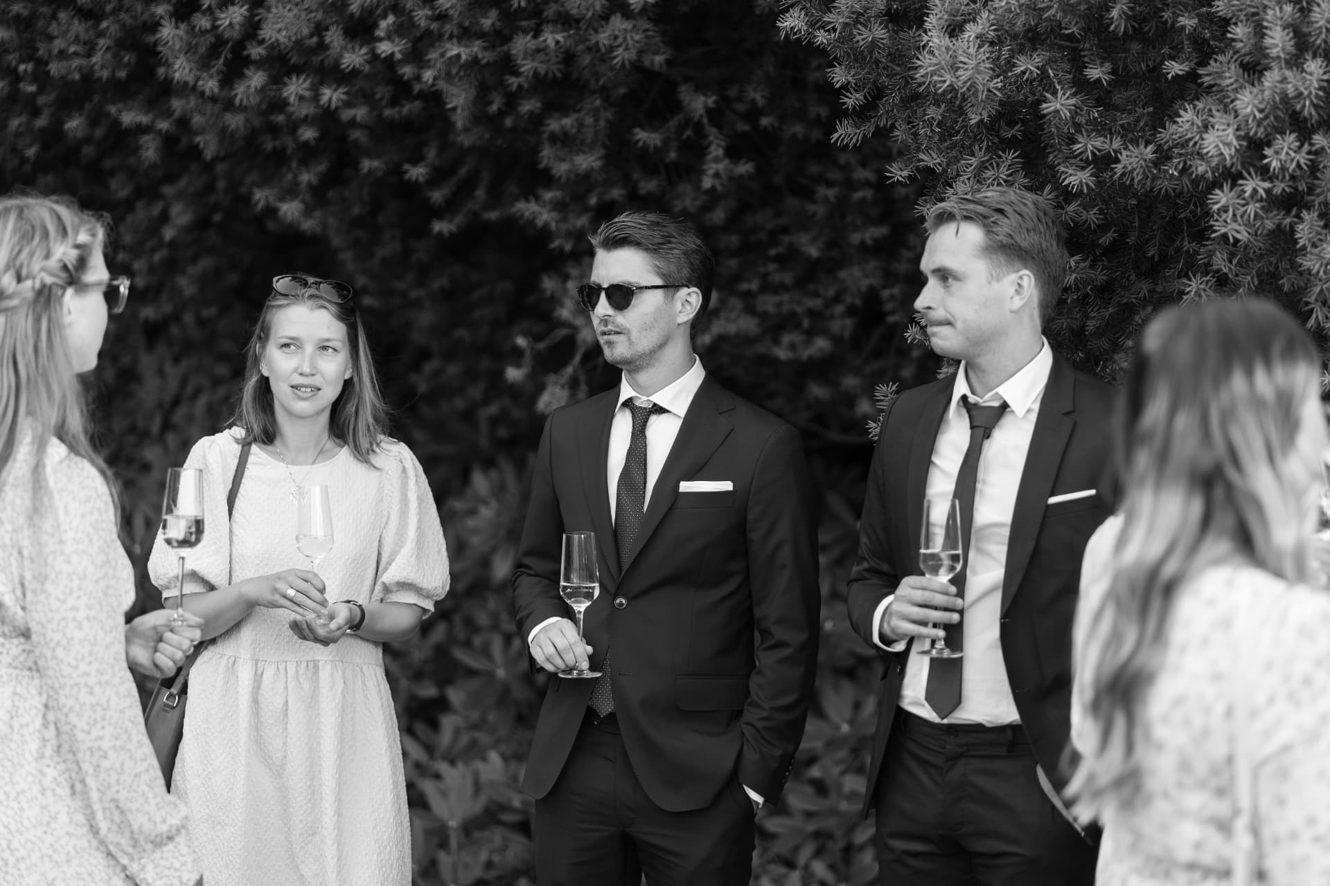 Gäster med champagneglas framför den mörka häcken i trädgården, svartvitt – bröllopsfoto av Mehmet Budak