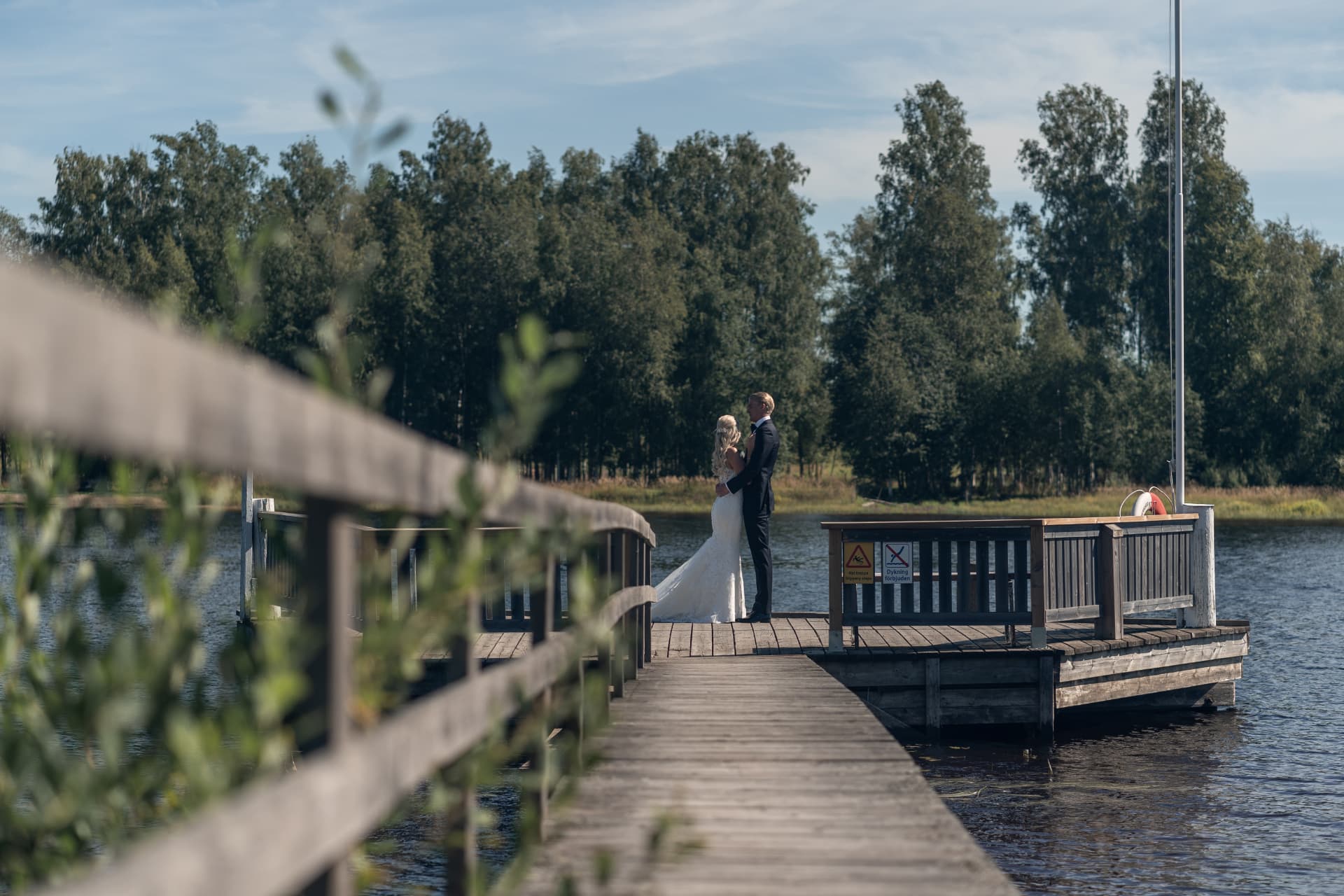 Brudparet står tillsammans på bryggan sett genom räcket vid sjön – bröllopsfoto av Mehmet Budak
