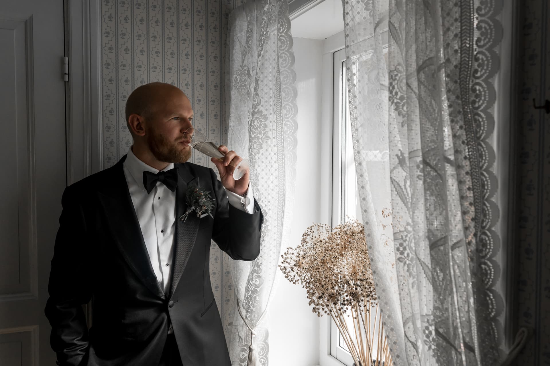 Brudgummen skålar vid fönstret med spetsgardiner – bröllopsfoto av Mehmet Budak